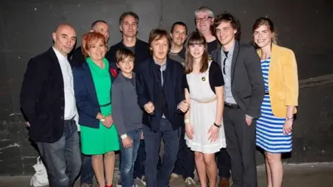 Eddie Janssens A group of 10 men, women and children stand smiling. Paul McCartney stands in the middle of the group. 