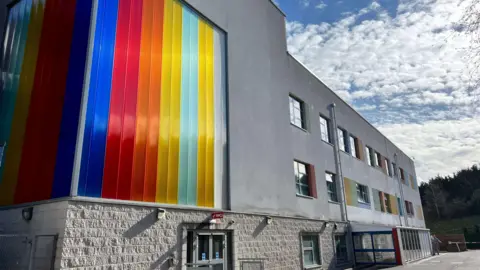 A grey hospital building with a window covered in a multi-coloured rainbow panelling. 