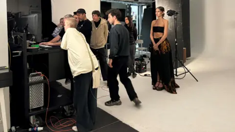 Family handout Liz looking at a computer screen with Ranking and five other people. There is a model in the background wearing a black long flowing skirt and a black bandeau top