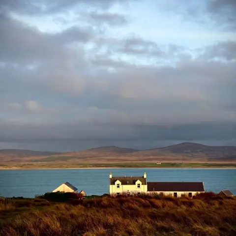 Cathy Cassie A white-walled two-storey house with a single storey extension attached sites on the shores of the loch. There are low hills in on the opposite shore.