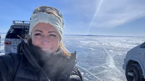 Anya smiles at the camera in a selfie. She is wearing a thick black coat and scarf, and a white knitted headband. There are a couple of 4x4 vehicles behind her, on a huge expanse of frozen lake. It is very sunny, with blue skies. 