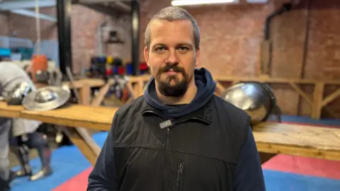 A man with short hair and a beard looks into the camera and smiles. Behind him, armour and a helmet rest on the edge of a wooden ring.