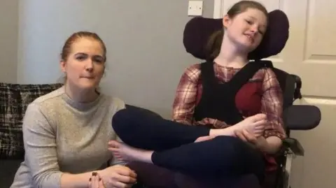Claire McGill is sat beside her daughter Sianon, who is in a wheelchair. 
Both are in a living room. Claire has her hair pulled back and is wearing a grey sweatshirt.
