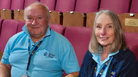Martin Giles/BBC Peter Bell and Susie Bell sitting on seats inside Norwich Theatre Royal. Peter is on the left in a blue polo shirt and is looking directly at the camera and smiling. Susie is sitting looking directly at the camera and smiling and is wearing a blue polo shirt and navy fleece.