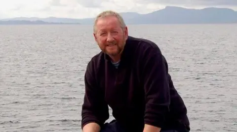 Alan Beattie Herriot Alan Beattie Herriot duduk di atas batu dengan laut di belakangnya. Dia memiliki rambut pendek dan janggut.