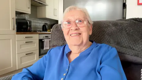 An elderly woman with short grey hair, clear framed. glasses and a blue shirt sitting on an armchair in a kitchen 