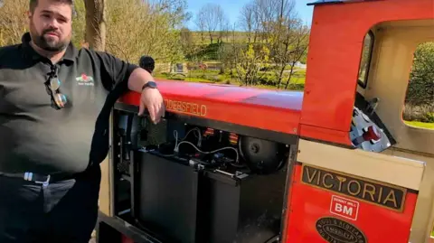 A man in a black top and trousers next to a small train with the word Victoria on the side.