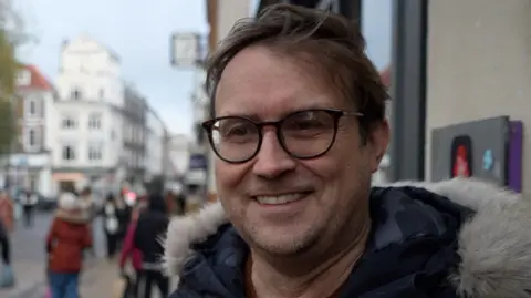 Qays Najm/BBC David Jackson tiene el pelo corto y castaño y lleva gafas de montura negra. Lleva una chaqueta azul oscuro y está parado frente a un edificio. Tiene una amplia sonrisa.