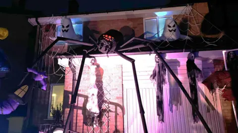 Photo shows a giant black spider with red eyes attached to a detached house. The spider's legs span from the floor to the roof of the garage. Spider's web and inflatable ghosts are also displayed around the spider.