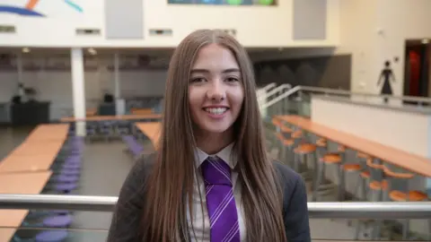 Hannah Karpel/BBC A teenager with long straight dark hair smiles at the camera. She is wearing a dark school blazer and a purple tie. 