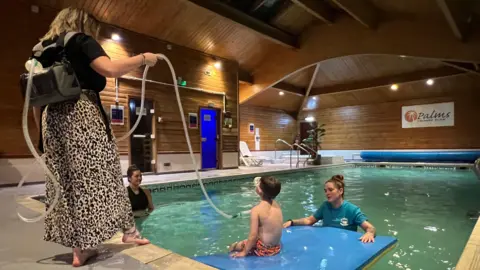 Henry sits on a float in a swimming pool, supported by Kayleigh and watched by Shevonne. A woman stands at the side of the pool. She is dressed in a black top and a long, leopard-print skirt and wears a black backpack containing a ventilator. She holds a long tube, which is attached to the backpack and runs to Henry, where it is attached to his neck.