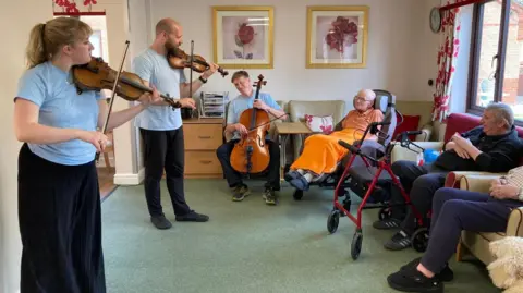 A cellist and two violinists perform in a nursing home day room while residents look on.