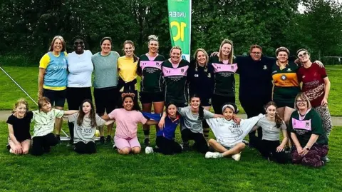 Whitehall RFC Juniors A group of female rugby players stand in a team shot facing the camera at Whitehall Rugby Club in Bristol. The front line is all young players and the more senior players are at the back with their arms linked