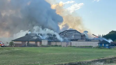 A golf club building with boarded up windows with smoke billowing upwards and part of the roof destroyed.