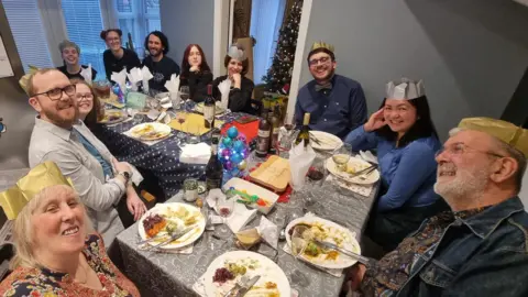 Ingrid Lyon Eleven people sitting around an L-shaped table, which is covered with a cloth and has plates of Christmas dinner placed in front of all the diners. Seven are wearing paper gold-coloured crowns. There is a decorated Christmas tree in the corner. All are smiling at the camera.