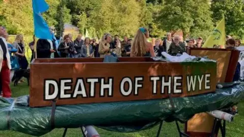 Campaigners gather on a river bank with a coffin which says Death of the Wye on its side. You can see grass below and trees in background.