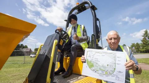 Councillor Bhupinder Gakhal sit atop a yellow digger with one hand on the steering wheel and one hand with a thumbs up. Standing on a green field below  who is wearing a yellow hi-vis jacket and is holding up a map of the field.