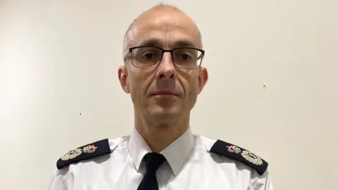 Mariam Issimdar/BBC Paul Sandford standing in front of a white wall inside BBC Radio Norfolk. He is looking directly at the camera and is wearing his police uniform of a white shirt and black tie.