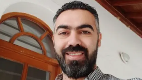 Amr Shayah An image of Amr, who has a black beard is smiling. He is wearing a black and white checked shirt and is standing in front of a white wall with an arched wooden frame.