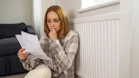 Woman sits with back to a radiator looking at a bill.