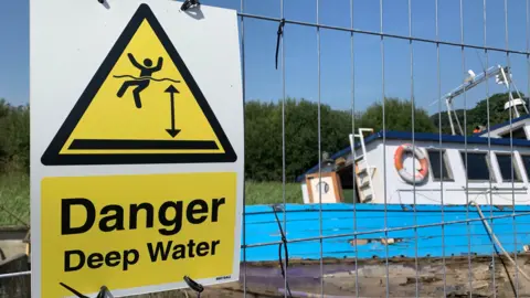BBC A danger deep water sign next to the wreck of a boat tipped on its side