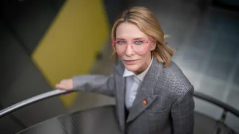 Tony Jolliffe/BBC News Cate Blanchett, wearing thin pink rimmed glasses is standing on a grey metal spiral staircase looking up at the camera. She has a grey checked jacket with a white shirt. Behind her, out of focus, in the basement is the yellow door to the Millennium Seed Bank. 