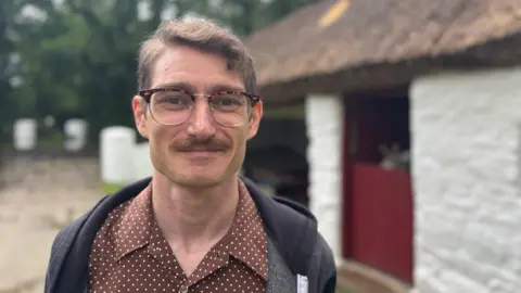 A man stands in front of a thatched cottage. He is smiling and dressed in a brown polka dot shirt with a hoodie over the top. He has short hair, a full mustache and is wearing tortoise shell glasses. Behind him is a whitewashed cottage and stable. A red door is half open with some donkey ears peeking out.