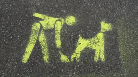 Stencilled image sprayed in yellow paint on pavement depicting a person bending down to scoop up a dog's poo