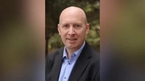 BT Group A portrait photo of Peter Bell. He is smiling at the camera. He is bald and wears a navy suit jacket with a light blue shirt underneath. Trees and bushes are behind him. 