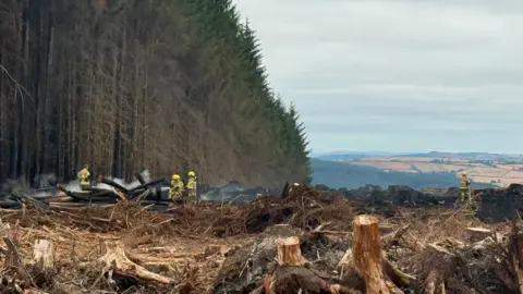 Market Drayton Fire Station Smoke rises from charred piles of wood next to a forested area at the top of a hill. Fields can be seen in the far distance and firefighters in yellow uniform stand in the near background.