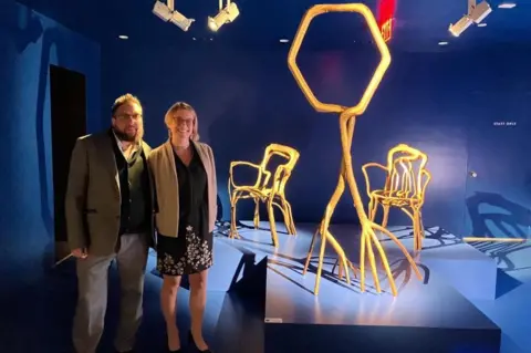 Supplied A man and a woman stood in front of two chairs made out of trees in an exhibition hall.