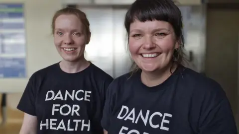 Dance for Health Two women, wearing the same black T-shirt, smiling and looking straight at the camera. The woman on the right has dark hair, in a ponytail and the other woman has auburn hair, in a ponytail. 