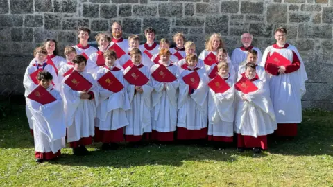 Truro Cathedral Choir A group of boys and girls wearing choir cassocks and surplices stand outside a granite church on the Isles of Scilly 