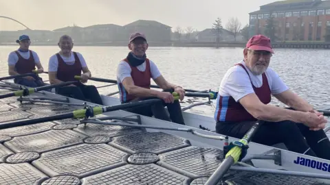 Four men with white, red and blue tops, three with caps, sitting in a rowing boat on the water. Buildings can be seen behind them.