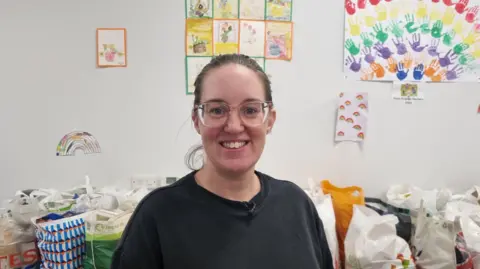 Carlisle foodbank's operations director Steph Humes inside the foodbank. She has brown hair tied back and wears glasses. She is wearing a black t-shirt and is smiling at the camera. Behind her are supermarket-branded bags filled with food. On the wall there are children's drawings and paintings.