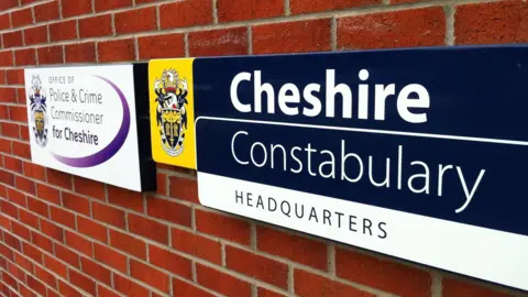 A sign with the words 'Cheshire Constabulary Headquarters' is positioned on a red brick wall. To the left is a second sign reading 'Office of Police & Crime Commissioner for Cheshire'