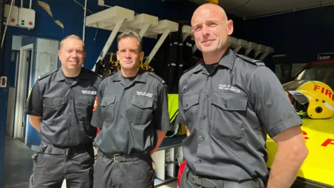 Jonny, Nick and Chris are wearing Jersey Fire and Rescue Service uniforms. They all look at the camera with their hands behind their backs. They are standing in front of a 4x4 fire and rescue vehicle.