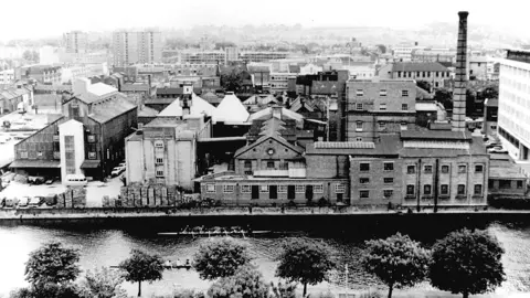 Charles Wells An old black and white image of a brewery, with a large chimney to the right. Other buildings are around it, with a river at the front, with trees along one bank. High-rise buildings are in the distance. 