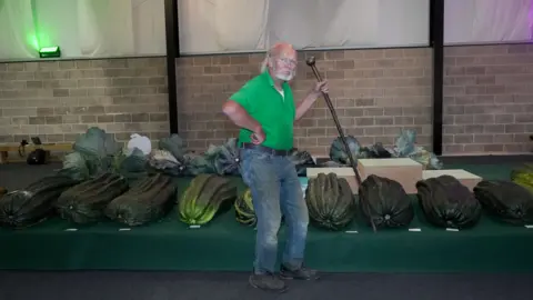 A man with a walking stick, held in the air, hand on his hip, standing in front of a line of huge marrows.