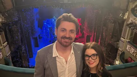 Samuel Moore Samuel Moore stands on the balcony of the Royal Albert Hall with his partner. He is facing away from the stage, which is lit in blue, white and red blocks. He is wearing a grey blazer and a white shirt. His partner is wearing black rimmed glasses, a black jumper and red lipstick.