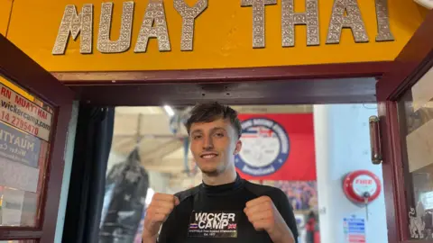 Simon Thake/BBC A young man in a black sports shirt raises his fists in a boxing stance. Above it, in large silver letters on a gold background, are the words: "Muay Thai".