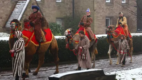 Three camels with red cloths covering their backs and colourful bridles, being ridden by three people dressed as wise men, led by two men and a woman in striped robes. They are walking on a snowy path past brick dwellings.