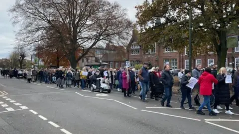 A line of people marching along one side of a road. Some are carrying a piece of paper or a placard.