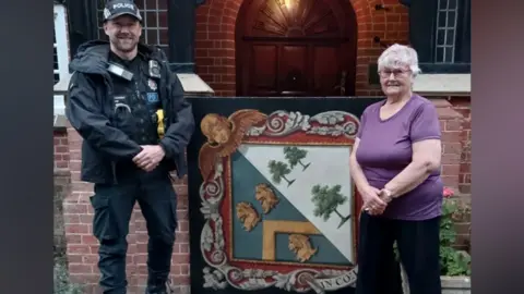 Essex Police PC Dane Wyatt, in full black police uniform, with a cap on, standing next to a large painted funeral hatchment, with Jan Nightingale, the other side. She is wearing a purple top, black trousers, with her arms crossed in front of her. They are by a building with a large wooden door. They are both smiling and looking at the camera. 