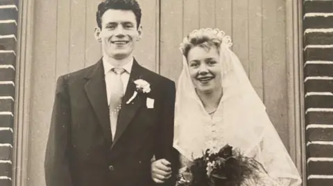 Family photograph A black and white image of a bride and groom, Brian and Isobel Teasdale, in front of a church. She is wearing a veil and is holding a bunch of flowers and foliage. He has on a suit with a carnation in his buttonhole. Both are smiling.