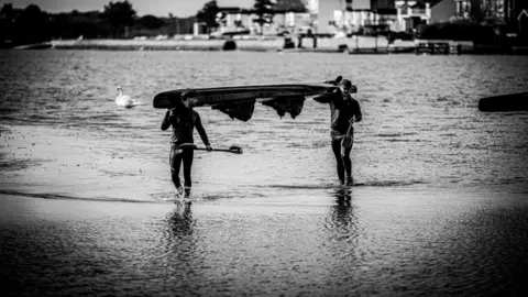 Mission Memory Ben Watkins and Oliver Smee carry a boat out of the water. They are in wetsuits and the picture is in black and white.