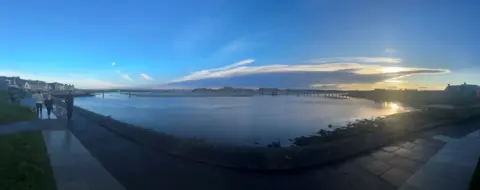 Pete Wilson A panoramic photo of a harbour with a blue sky above and three people walking at the left-hand side