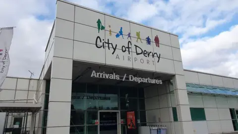 BBC Front of City of Derry Airport building. It has graphics of people above the City of Derry Airport sign on a white building. There is an Arrivals/Departures sign above a glass entrance door.