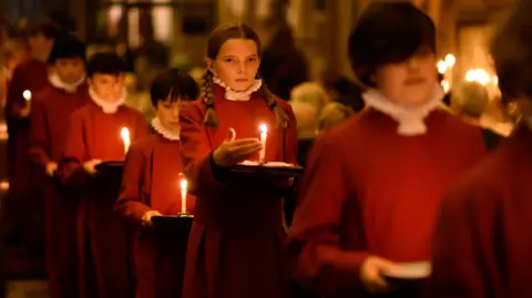 Jon Craig Young choristers walking down the aisle of Bristol Cathedral, dressed in red robes and white neck cuffs, all holding candles in the darkness