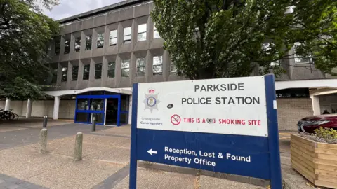 The grey concrete exterior of Parkside Police Station. A sign identifying the location, on a blue backing, stands in front of the building.